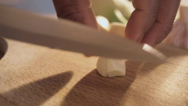Macro shot of male African American hands chopping a clove of garlic into thin pieces with a sharp chef�s knife on a wooden board.