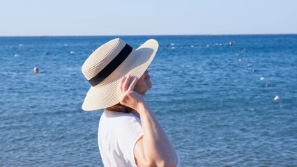 Back view of a woman in a straw hat and white dress looking at the blue sea, close up. The concept of relaxation, rest, sunscreen