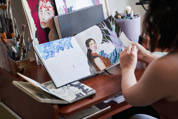Female artist sitting at the table in her art studio and looking at her art works