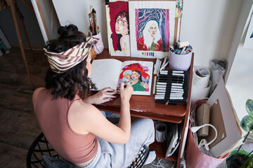 Female artist sitting at table wand making some illustrations with color markets at home