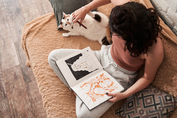 Female artist playing with cat while drawing at her notebook from home