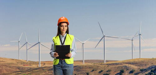emale engineer with a safety vest and hardhat holding a clipboard on a wind turbine field © Ljupco Smokovski