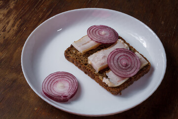 whole grain rye bread and prickled pork lard sandwich with red onions on white plate, wooden table....