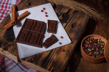 Broken bar of dark bitter chocolate with chilly pepper, cinnamon, red pea pepper inside on paper,...