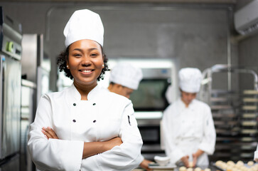Selective focus of an African female baker in a chef uniforms, standing with arms folded and smiling at camera, with blurred colleagues kneading the dough in the background. Copy space on right side. - Powered by Adobe