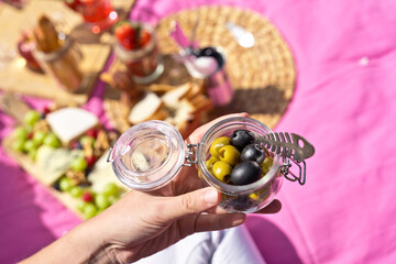 Horizontal top view of unrecognizable woman holding a bowl of olives outdoors. Above shot of woman holding black and green olives on pink picnic background. Health and food concept.
