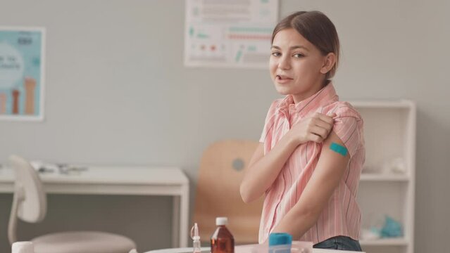 Medium slowmo portrait of smiling 13-year-old Caucasian girl sitting on medical couch at doctors office looking at camera showing her shoulder with band aid after vaccine injection