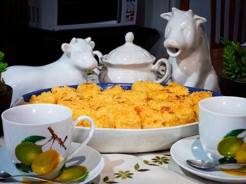 Pieces of cake on a serving bowl, white teapot and butter dish in the shape of a cow, two white tea cups with lemmon painting on a table with green floral  embroidery . Microwave behind, kitchen.