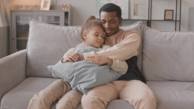 Medium slowmo of African American man and his pretty 8 year old daughter sitting together on couch embracing each other, staying at home during lockdown