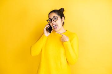 Young beautiful brunette woman wearing glasses having conversation talking on smartphone very happy pointing with hand and finger