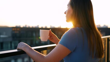 Woman with a cup of coffee standing on the balcony and admire the sunset. Slow motion