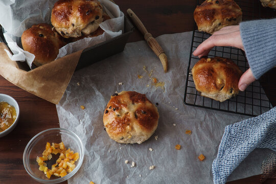 Traditional Easter food image of freshly baked hot cross buns. Baking equipment and ingredients included in frame. Woman's hand entering right side of frame to take a glazed bun. 