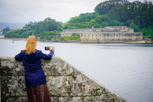 blonde young woman with blue coat taking photos at Castillo de San Felipe. Stone and earth coastal castle from the 16th century with views of the Ferrol estuary