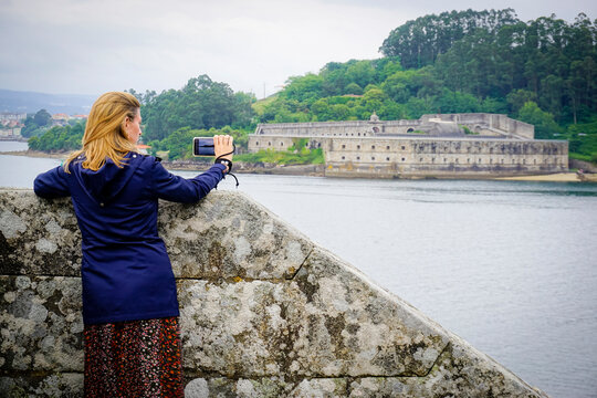 blonde young woman with blue coat taking photos at Castillo de San Felipe. Stone and earth coastal castle from the 16th century with views of the Ferrol estuary