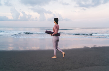 Slim female yogi dressed in trendy sportive wear holding violet mat choosing place for morning training at sandy beach, Caucasian woman in tracksuit walking at seashore coastline during weekend