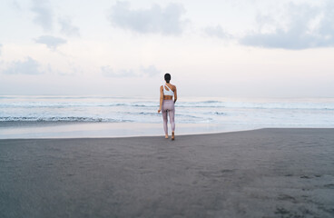 Back view of slim female yogi in stylish tracksuit walking to ocean water during morning time for training at coastline, good looking woman in sportive wear have promenade at seashore beach