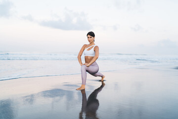 Portrait of flexible female trainer in sportive tracksuit looking at camera while stretching body muscles and doing warm up exercises at coastline sand, healthy woman keeping vitality and wellness