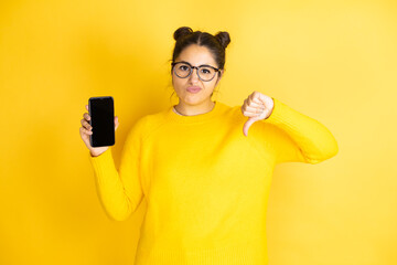 Young beautiful brunette woman wearing glasses holding smartphone showing screen with angry face,...