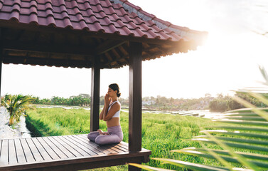 Side view of tranquil female in namaste praying near Indonesian rice fields and meditating for get mindfulness and enlightenment, calm woman in sportswear enjoying morning yoga practice in Thailand