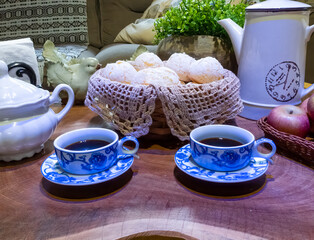 Brazilian cheese bread in a basket, red apples in a basket, coffee cups, coffee pot, sugar pot and potted plant on a table. Chair, sofa and cushions behind