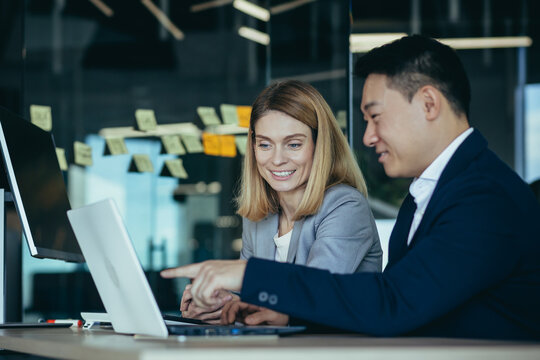 Happy Coworkers Discuss Project Strategy By Looking And Pointing At Laptop Pc Computer Monitor Screen. Multiethnic Business Team In The Office. Confident Mature Asian Man Explaining Young Female