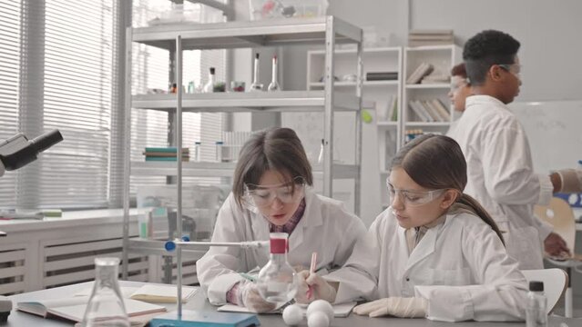 Medium slowmo of two Caucasian schoolgirls in lab coats and protective glasses doing pair lab work at Science class