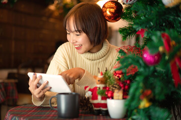 Beautiful woman in sweater smiling and sitting on chair to chatting with friend on tablet near christmas tree