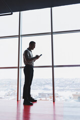 A young executive is holding a phone while standing in an office interior and looking out a large window overlooking the city. Male manager with a phone is thinking about a new business project