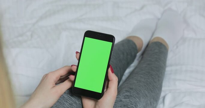 Woman sitting on the bed and using smartphone with pre-keyed green screen. Female tapping at the screen. Mock up and chroma key concept. Close up
