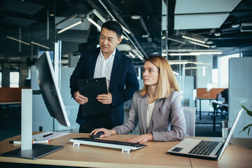 Two Asian male and female colleagues in a modern office, a woman shows the work done on the monitor, consults and discusses - Powered by Adobe