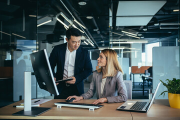 Two Asian male and female colleagues in a modern office, a woman shows the work done on the monitor, consults and discusses