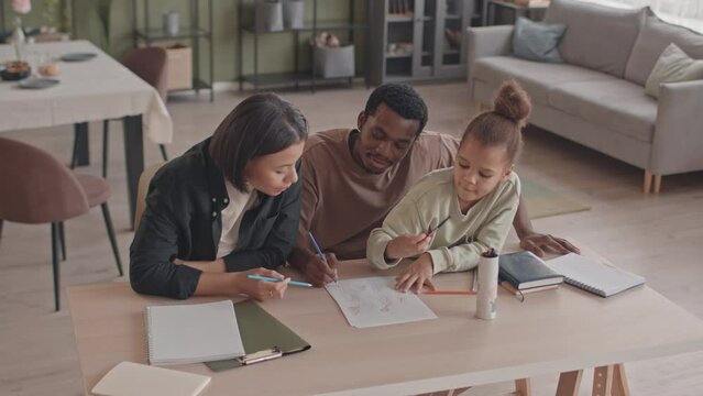 From above slowmo of little African American girl and her parents sitting at desk in cozy living room drawing pictures together