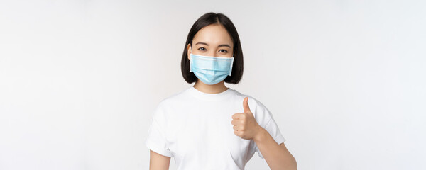 Smiling asian girl in medical mask showing thumbs up, approve something good, praise and compliment...