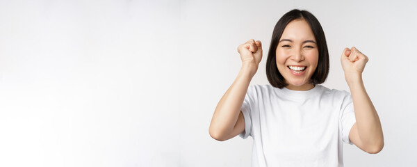 Naklejka na ściany i meble Portrait of enthusiastic asian woman winning, celebrating and triumphing, raising hands up, achieve goal or success, standing over white background