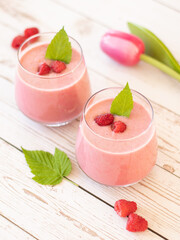 Pink raspberry smoothie in a glass on a wooden table with fresh leaves and raspberries. Healthy fruit drink for breakfast or dessert. Vegan plant-based food and beverage concept.