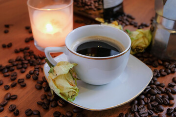 A coffee cup with a yellow rose on the side and a lighting candle in the background.