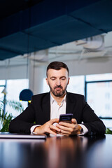 Confident male businessman in a business suit sits at a table with a phone and works remotely