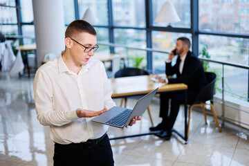 Fototapeta na wymiar A young businessman man in glasses and a white shirt with a laptop in his hands stands in the office and works