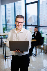 Fototapeta premium A young businessman man in glasses and a white shirt with a laptop in his hands stands in the office and works