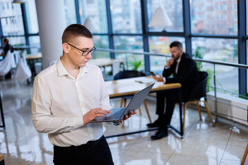 A young businessman man in glasses and a white shirt with a laptop in his hands stands in the office and works