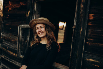 Obraz na płótnie Canvas beautiful Ukrainian girl in black clothes and a straw hat stands near the old wooden house. The war in Ukraine. Portrait of a woman on a dark wooden background. Flee the war to the village