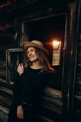 beautiful Ukrainian girl in black clothes and a straw hat stands near the old wooden house. The war in Ukraine. Portrait of a woman on a dark wooden background. Flee the war to the village