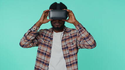 Exited african american man using virtual reality futuristic technology VR app headset helmet to play simulation 3D 360 video drawing game. Young guy on blue studio background. People sincere emotions