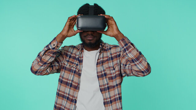 Exited african american man using virtual reality futuristic technology VR app headset helmet to play simulation 3D 360 video drawing game. Young guy on blue studio background. People sincere emotions