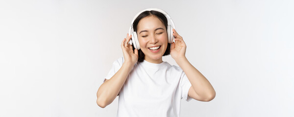Fototapeta na wymiar Modern asian girl dancing, listening music with headphones, smiling happy, standing in tshirt over white background