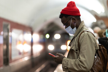 Young African American hipster man wear face mask in subway using smartphone waiting for train on...