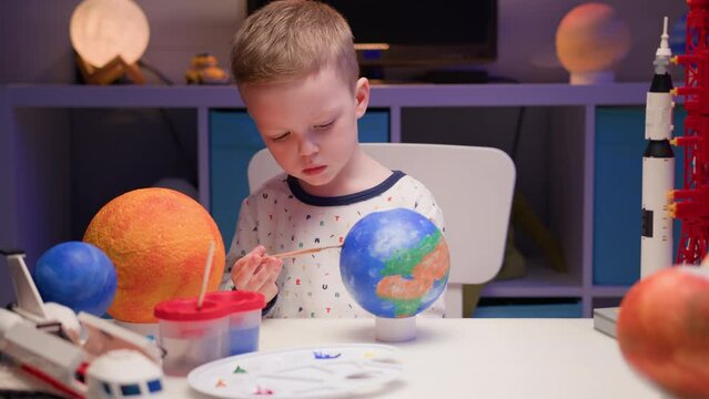 Blond boy paint planet solar system Earth with colorful paint sitting home table in evening, planet solar system, spaceships and space shuttle from constructor around. Cosmonautics Day on April 12.