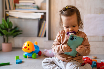 One baby small caucasian infant girl playing on the floor at home copy space holding mobile phone smartphone using to call or watch video in day front view