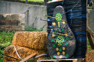 Traditional arrow target board with arrows stuck on it near haystack on farm.  Hay bales in a...