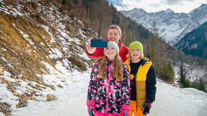 Joyful family, the father with daughter and son, taking selfies with a smartphone in front of a winter mountain scenic landscape.